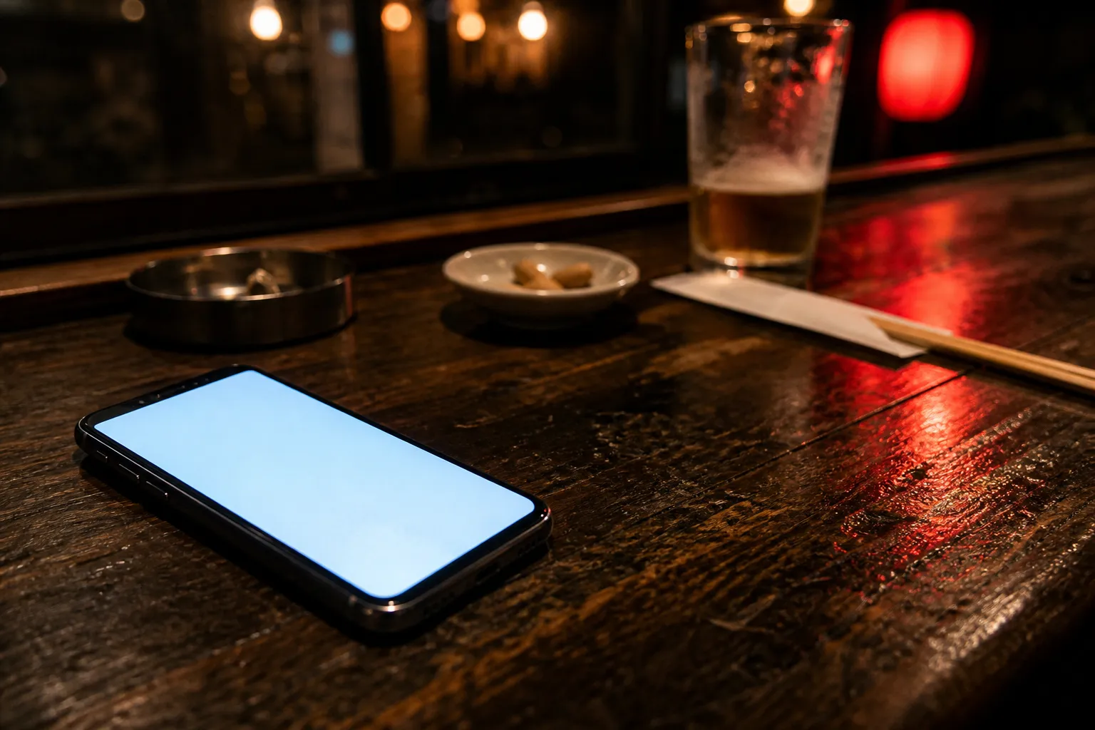 Phone glowing face up on a wooden izakaya counter at night beside a half-finished beer